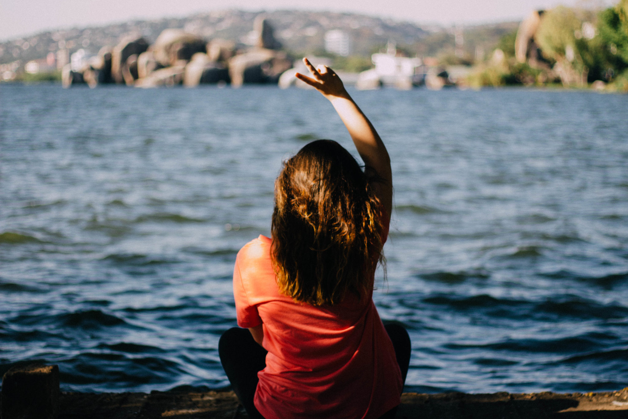 A student sits by the water with her back to the camera holding up her fingers in a peace sign.