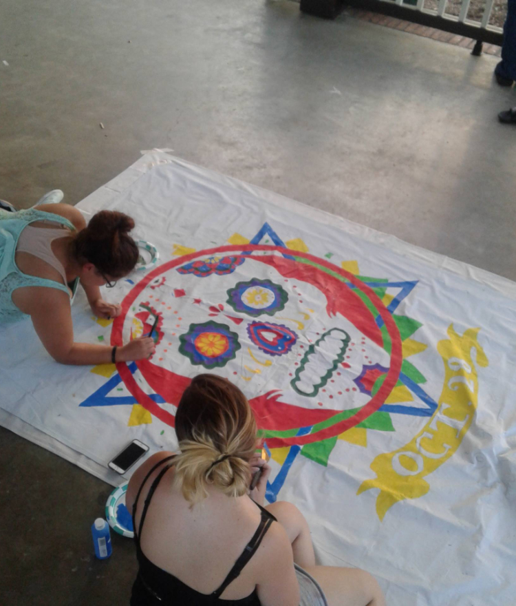 two students paint a decorated calavera on a white sheet