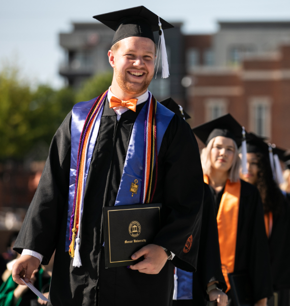 a student wearing a cap, gown and cords carries his diploma under his arm