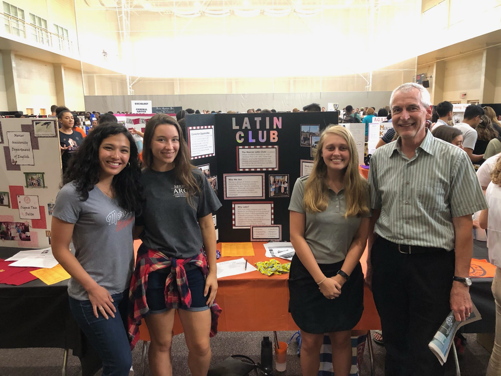 three students and a professor stand smiling in front of a trifold board that says latin club