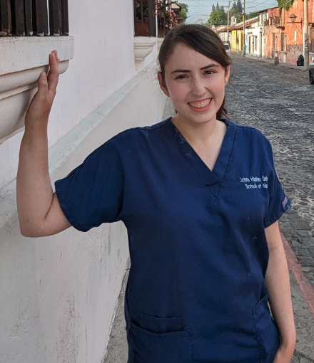 A smiling Brianna Levin wears blue medical scrubs while standing on a cobblestone street, resting an arm on a white wall, with buildings in the background.