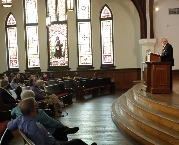 A group of people is seated in wooden pews inside Willingham Hall, with ornate stained glass windows in the background. They are listening to a speaker who is standing at a podium on a raised platform.