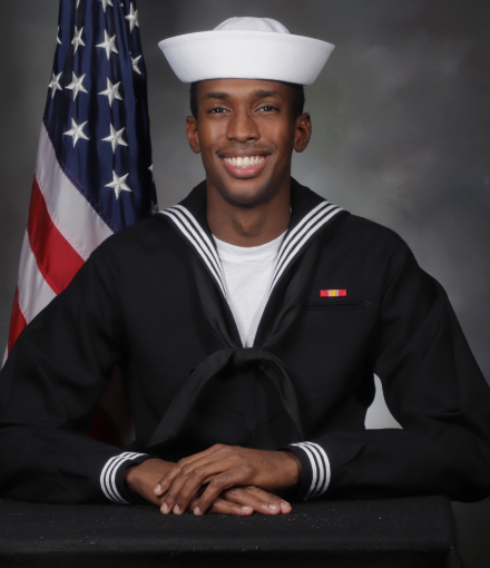 An individual wearing a U.S. Navy uniform sits in front of an American flag.