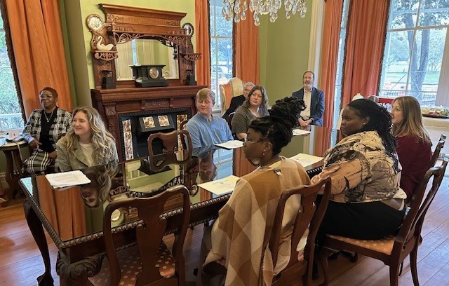 A group of people is seated around a large wooden table in an elegant, historic-looking room with green walls and large windows with orange curtains. The room features a decorative wooden fireplace mantel and a chandelier. The group appears to be engaged in a meeting or discussion, with papers and notebooks in front of them.