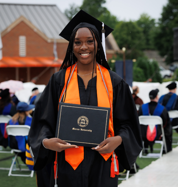 A Mercer graduate wears a black graduation gown and cap and an orange stole. She is holding a Mercer degree. Commencement exercises continue in the background.