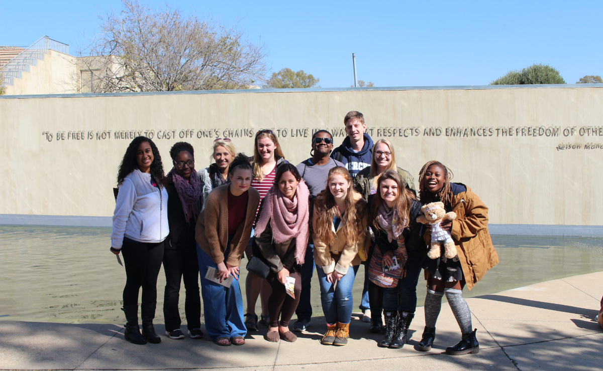 Twelve Mercer students pose for a group photo in front of a wall that features this quote from Nelson Mandela, To be free is not merely to cast off one's chains but to live in a way that respects and enhances the freedom of others.