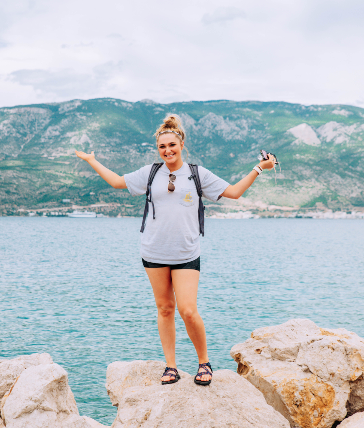 A Mercer student stands on a rock in front of a turquoise body of water. Lush mountains are behind her.