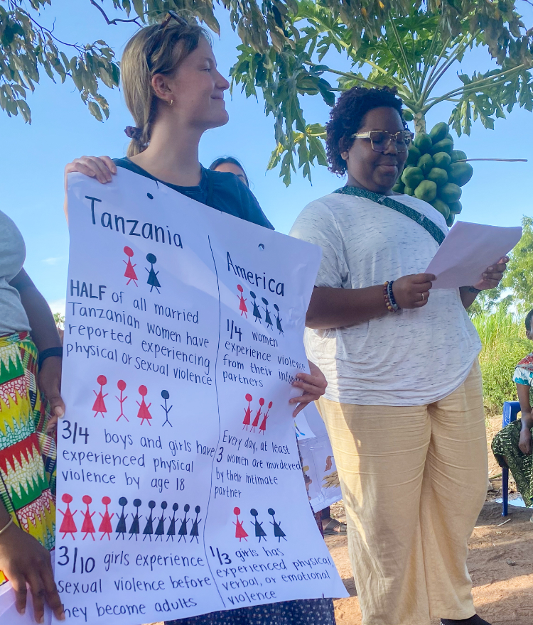 A Mercer student holds a poster featuring red and black stick figures that compares violence against girls and women between Tanzania and the U.S. Another student stands next to her looking down at a paper.