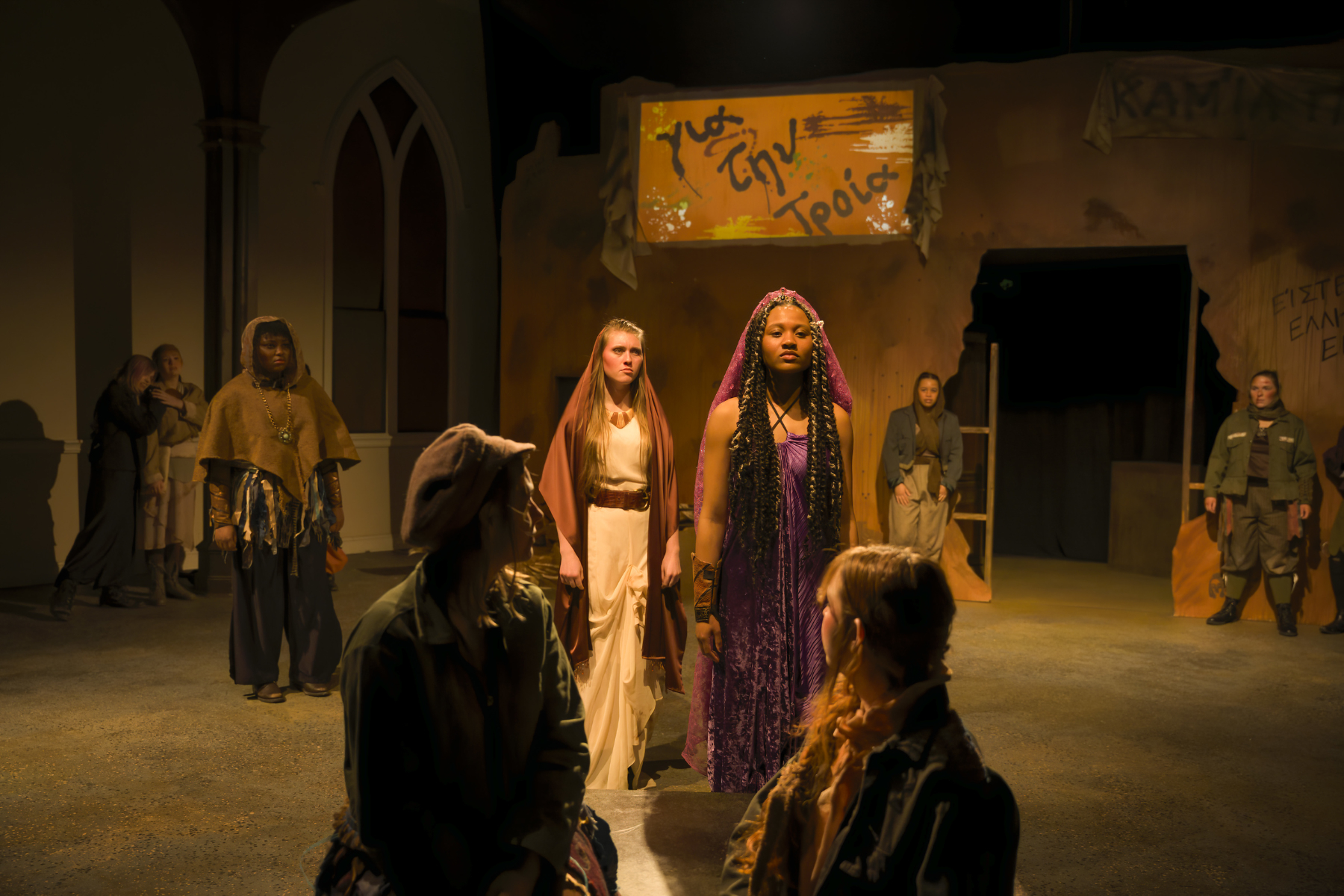 Actors in a dramatic stage scene stand under warm lighting, with a woman in purple at the center and others in muted costumes.