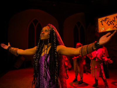A Mercer Theatre actor in a purple costume stretches her arms wide during a dramatic scene in Trojan Women, bathed in red stage lighting.