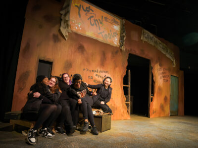 Five Mercer Theatre cast members in black costumes laugh and embrace on stage in front of a graffiti-covered set for Trojan Women.