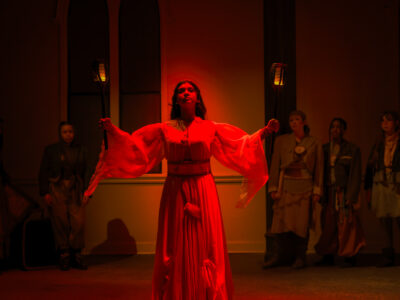 A Mercer Theatre actor in a flowing red gown holds two torches aloft center stage during a dramatic scene in Trojan Women, bathed in deep red lighting.