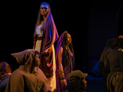 A Mercer Theatre actor in a draped robe stands elevated above the cast during a solemn scene in Trojan Women, lit in warm gold and blue.