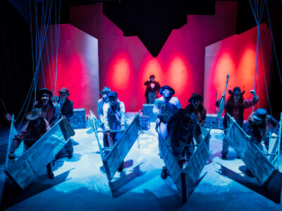 Mercer Theatre cast performs Men on Boats, rowing prop boats on a dramatic stage lit in blue and red with ropes hanging overhead.