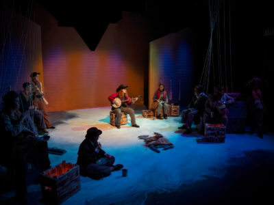 A Mercer Theatre actor plays banjo at center stage while the cast of Men on Boats rests around her in warm and blue stage lighting.