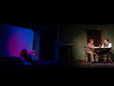 A split stage in Grand Guignol shows one actor huddled alone in blue and pink light while two others confer at a table in a separate lit room.