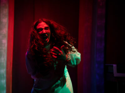 A Mercer Theatre actor screams with wild hair and outstretched hands during an intense horror scene in Grand Guignol, bathed in deep red light.