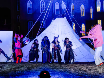 Mercer Theatre actors in elaborate fantasy costumes with horns and dark armor perform an outdoor night scene in Dragon's Dream in front of a lit campus building.
