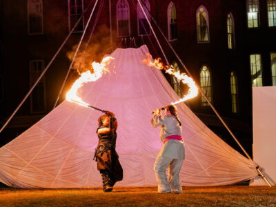Two Mercer Theatre actors wield flaming swords in a dramatic duel during an outdoor night performance of Dragon's Dream in front of a glowing campus building.