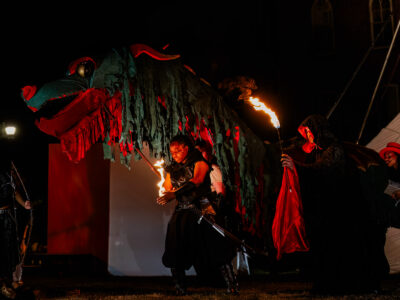 Mercer Theatre actors in fantasy costumes wield fire beside a massive dragon puppet during a dramatic outdoor night scene in Dragon's Dream.