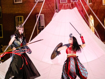 Two Mercer Theatre actors in elaborate fantasy armor and face paint clash with spears and a shield during an outdoor night battle scene in Dragon's Dream.
